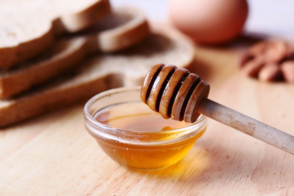 Close-up of honey dipper in a bowl with honey on wooden table beside sliced bread.